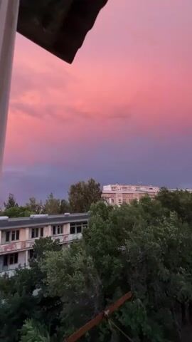 This video captures a breathtaking sunset over Tokmok City, Kyrgyzstan. The sky is a vibrant spectacle of pink and purple clouds, transitioning from a soft rosy hue at the top to a deep, moody indigo lower down. The vibrant colors suggest the presence of high-altitude clouds catching the last rays of the sun.

In the foreground, a dark, angular object, possibly a rooftop or balcony railing, partially obscures the view, adding a sense of depth. Below the dramatic sky, a cityscape unfolds. Several buildings are visible, some appearing to be older, with visible wear and tear on their facades, while others are more modern, with light-colored walls. Lush green trees fill the middle ground, creating a natural barrier between the buildings and the viewer, their foliage dense and verdant.

Further into the distance, a parking lot is visible, with a few cars scattered across it, hinting at the ongoing life of the city even as the day ends. The overall scene evokes a sense of peaceful transition from day to night, with the dramatic sky providing a powerful emotional anchor. The time of day is clearly sunset, and the weather appears calm, with the vibrant colors indicating fair conditions conducive to such a display. There is no visible text in the video.