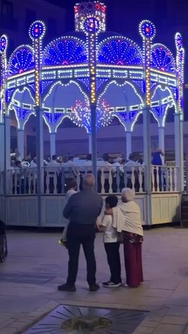 A family of four, a man, a woman, and two boys, stands facing a brightly lit gazebo. The gazebo is decorated with numerous blue and white lights, with some red lights at the very top. A band appears to be performing on the gazebo. The family is standing on a paved area in front of the gazebo. The lighting suggests it is nighttime.