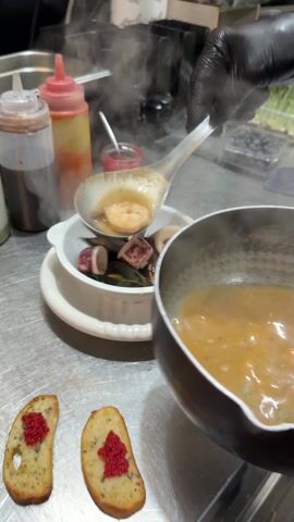 A cook wearing black gloves prepares a seafood dish in a commercial kitchen. The cook uses a large ladle to transfer steaming, tan-colored broth from a silver pot into a white bowl containing mussels, shrimp, and calamari.  On the stainless steel countertop, next to the bowl, are two slices of toasted bread topped with a dollop of bright red caviar-like beads. In the background are several squeeze bottles of sauce and a container of blueberries. There is steam rising from the dish and pot.