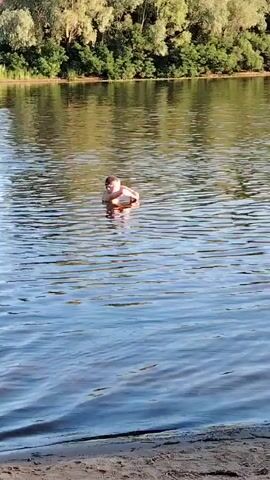 A person is swimming in a body of water. The person is visible from the waist up and appears to be treading water. The water is calm and reflects the trees and sky in the background. The person is wearing dark shorts and is facing the camera. The person then submerges into the water.