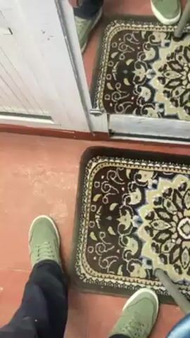 A person stands in front of a mirror. The person wears black pants and light green shoes, and stands on a brown floor with a decorative rug in the foreground. As the camera rises, the reflection in the mirror shows a man wearing a black, long-sleeved, zippered mock turtleneck. He holds a white cellphone in front of his face. Behind him, clothes are hung on a rack. The lighting appears to be bright, and the setting resembles an interior space.
