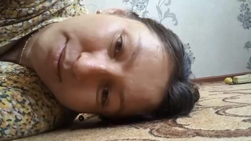 A low-angle, close-up shot shows the face of a person lying on the floor. The person's head is tilted to the right, with their hair falling to the left. They are wearing a floral patterned shirt. The background features a wall with a floral wallpaper and a wooden floor. A yellow object and a white object are visible on the floor in the background.