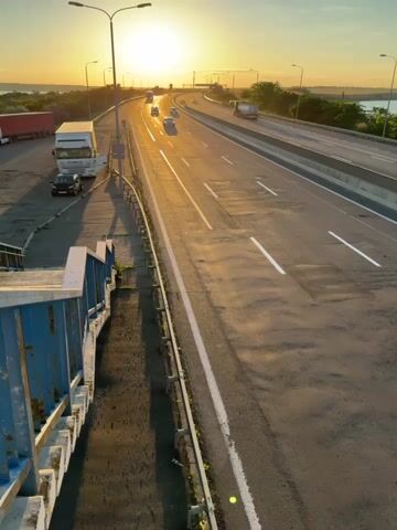 The video shows a wide, multi-lane highway at sunset. The sun is low on the horizon, casting a warm golden light across the scene and creating long shadows. Several vehicles are visible on the highway, including cars and trucks. On the left side of the highway, there is a pedestrian walkway with blue railings. The surrounding environment appears to be somewhat rural, with some vegetation visible beyond the highway and a body of water in the distance. The overall mood is peaceful and serene, characteristic of a sunset scene.
