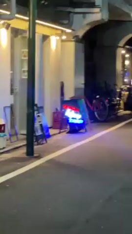 The camera slowly approaches a neon sign that reads “NOODLE BAR TENYUU” in red and blue. There are two other signs next to it, as well as a bicycle parked nearby. The scene appears to be in front of a restaurant or bar at night.