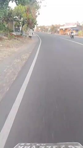 The video is shot from the perspective of someone riding a motorcycle on a paved road. The camera is pointed forward, showing the road stretching ahead. On the left side of the road, there is a sidewalk with trees and some structures that appear to be shops or small buildings. A white line marks the edge of the road. The sky is visible at the top of the frame, and it appears to be daytime, possibly late afternoon due to the lighting. There are a few other motorcycles visible in the distance on the road. The overall scene depicts a typical rural or suburban road in a tropical or subtropical climate.