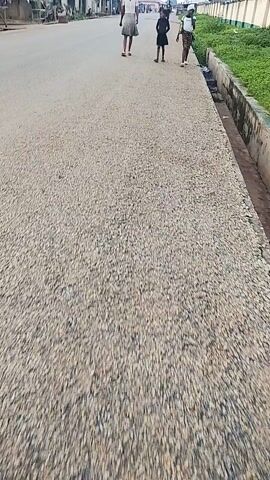 A person is filming from a slightly elevated, moving perspective, looking down at a gravelly, possibly unfinished road surface. Several individuals, including children, are walking on the road in the distance. The environment appears to be outdoors with some greenery visible on the left side of the frame, adjacent to a concrete structure. The sky is overcast. The footage is oriented sideways.