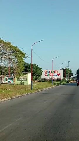 The video shows a view of a road with a large sign that says "Puerto Cabello está de Moda," meaning "Puerto Cabello is in Fashion." The sign is painted in bright, vibrant colors and features a red heart in the middle of the sign. The road is lined with trees and lamp posts that are painted red, white, and blue. A black SUV drives past the sign from left to right. The weather appears to be sunny with a clear blue sky.