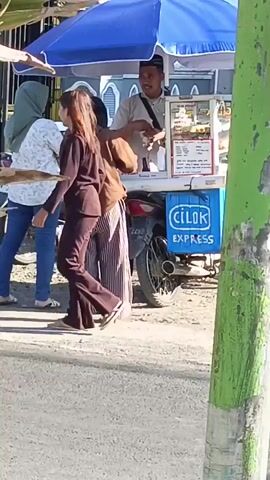 Two women and a man are at a food cart that is attached to a motorcycle. The cart has a blue umbrella and a sign that says "CILOK EXPRESS". One woman is wearing a brown hooded sweatshirt and striped pants. The other woman is wearing a white shirt with a floral pattern and blue jeans. The man is wearing a white shirt and a black hat and appears to be serving food from the cart. The background shows a fence and some vegetation. The ground is paved.