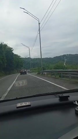 The video is shot from the perspective of a person in the passenger seat of a car. The car is driving on a paved road that curves to the left. There are other cars on the road, and the environment appears to be rural, with lush green trees and hills visible on either side of the road. The sky is overcast and gray. There are utility poles and power lines running alongside the road. The dashboard of the car is visible in the foreground. The video is quite shaky, suggesting the car is in motion. The time stamp at the beginning indicates the video is only a few seconds long.