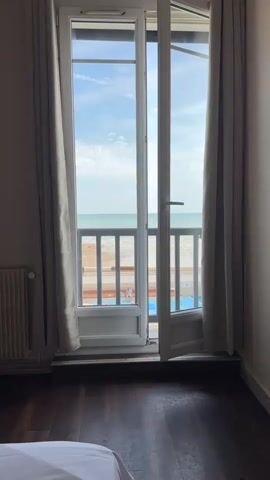 The video shows a view from a room looking out a large set of double glass doors onto a beach and the ocean. The doors are slightly ajar. The room has dark wooden floors and a white radiator on the left side. Beige curtains hang on either side of the doors. Outside, a wooden railing is visible in front of a sandy beach. A few people can be seen on the beach. The sky is blue with scattered clouds, and the ocean is a muted blue-green. The video appears to be a static shot with no movement.