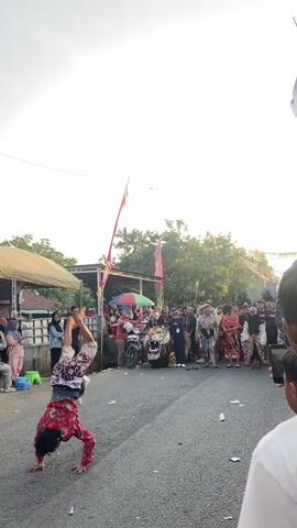 A man in a red patterned shirt and white shorts dances in the street. He is holding a pink and yellow flag on a stick. He is crouching and then stands up, raising the flag. He is surrounded by people watching him. There are also some motorcycles and a large tiger-like decoration in the background. There are flags and banners visible in the scene. The ground is asphalt. The sky is bright and overcast.
