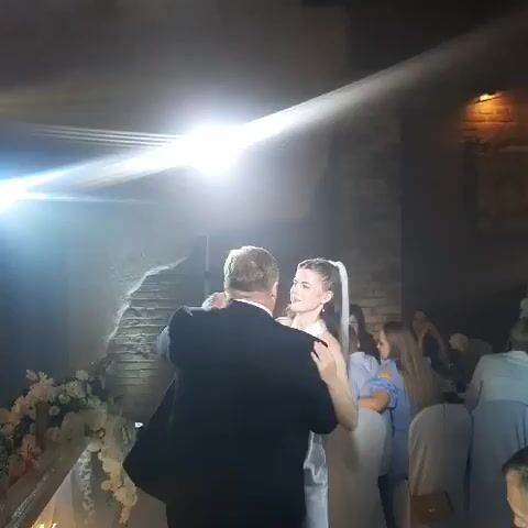 A man and a woman are dancing together at a wedding. The man is wearing a black suit and the woman is wearing a white wedding dress and veil. They are looking at each other and smiling. There are other people in the background, as well as decorations such as flowers. The lighting is dim with bright spotlights.