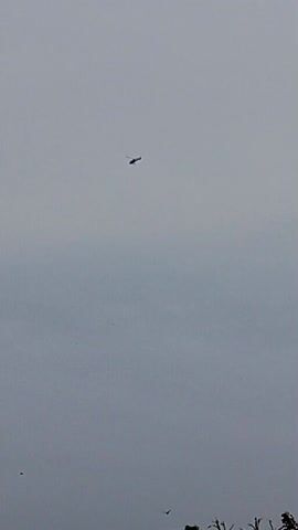 The video captures a distant helicopter flying across a uniformly gray, overcast sky. The helicopter, appearing as a small, dark silhouette, moves steadily from the right side of the frame towards the left. Its main rotor and tail rotor are discernible against the dull background as it passes. The distinct, rhythmic sound of helicopter rotor blades is clearly audible throughout the clip, subtly fading as the aircraft moves further away. The sky is entirely covered in clouds, creating a flat, diffused light, indicating daytime under overcast conditions. There are no ground-level geographical cues, people, buildings, or other vehicles visible in the frame that would specifically identify a location within Chah Sultan, Pakistan, beyond the provided context. No text is visible.