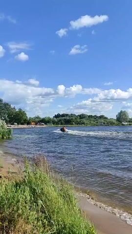 A jet ski with two individuals, one wearing an orange life vest, navigates a body of water, creating a white wake. In the background, a tree line borders the water, with several vehicles and people visible on the shore. The sky is blue with scattered white clouds.