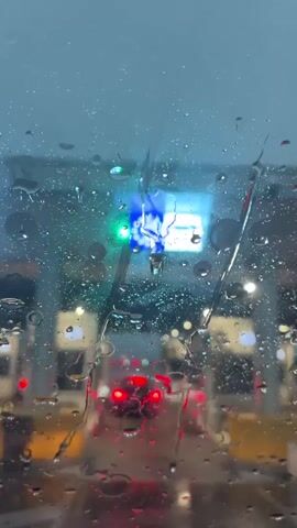 The video captures a first-person perspective from inside a car, looking through a rain-splattered windshield. Raindrops heavily obscure the view, streaming down the glass. Through the water, the red taillights of vehicles ahead are visible, appearing as blurred streaks of light.

Above the car lights, illuminated signs are discernible. A bright green light is visible on the left side of the frame. To its right, a blue illuminated sign features a white circle of stars, commonly associated with the European Union symbol. Adjacent to this, a white illuminated sign is also visible, though its details are unclear due to the rain and blur.

As the video progresses, the windshield wipers briefly clear a section of the glass, momentarily improving visibility of the signs and the structures ahead. These structures appear to be booths, consistent with a border crossing or toll plaza, with a car approaching them. The scene is illuminated by artificial lights, suggesting it is evening or night, and the weather is clearly rainy. The location context is Kalotina, Bulgaria, indicating a border crossing.