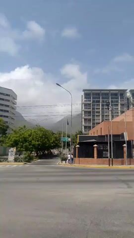 The video shows a street scene with several buildings, mountains in the background, and a cloudy sky. There are cars and a motorcycle on the road. In the foreground, there is a crosswalk with yellow lines. A white bus is driving past. The buildings are modern, with glass windows and balconies. The mountains are covered in greenery. The sky is blue with some white clouds. The overall impression is of a sunny day in a city with a mountainous backdrop.