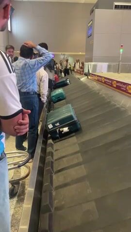 The video shows a baggage carousel in an airport. Several pieces of luggage, mostly teal and some red, are visible on the moving belt. A group of people are standing in a line along the edge of the carousel, waiting to collect their bags. Some individuals are looking at the carousel, while others appear to be talking on their phones. The environment is a typical airport baggage claim area, with overhead lighting and signage visible in the background. The perspective is from behind a person wearing a white and blue shirt, looking towards the carousel and the other passengers.