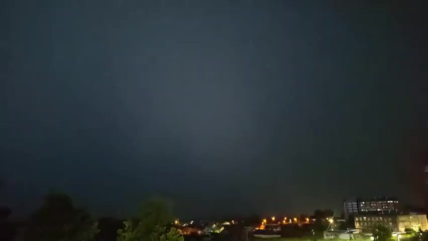 A wide shot captures a dark sky over a town at night. A massive lightning strike illuminates the sky with a bright white flash, briefly revealing silhouetted trees and buildings in the foreground and middle ground. The lightning strike is horizontal and spans almost the entire width of the frame. The sky then returns to darkness.