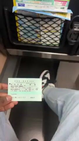 Here is a detailed description of the video:

The video begins inside a train, with a person holding up a train ticket for the camera. The ticket is green and has Japanese writing on it. The person holding the ticket is wearing jeans and sneakers. The train appears to be clean and well-maintained. The seats are black with a checkered pattern, and there are overhead lights. There is a net pocket on the seat in front of the person holding the ticket, with some leaflets inside. Then the camera pans to show the inside of the train car. There are overhead lights and a screen ahead, and the train appears to be mostly empty.