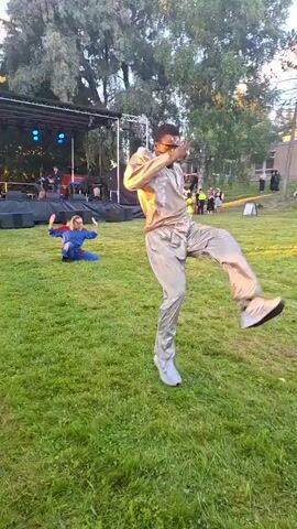 A group of people are performing martial arts on a grassy outdoor stage. In the foreground, a man in a light-colored martial arts uniform is performing a high kick. He is wearing white sneakers. Behind him, to the left, a person in a blue martial arts uniform is in a low stance. Further back, on the stage, a band is playing. There are trees and buildings in the background. The sun is setting, casting a warm glow on the scene.