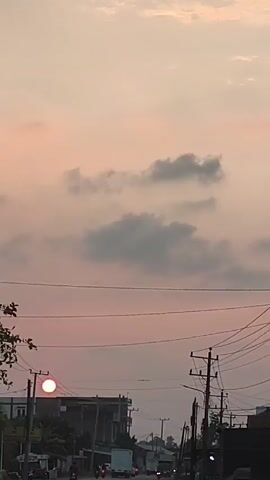 Here is a description of the video:

The video shows a sunset in an urban area. The sky is a mix of pink and gray, with some clouds visible. The sun is setting behind a building in the distance. There are power lines and poles in the foreground, and some trees on the left side of the frame.  There are some vehicles, including cars and motorcycles, driving on the road. There's also a storefront to the left. The video appears to be shot from a moving vehicle.