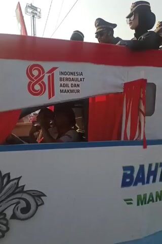 The video shows a parade float designed to resemble a ship. Several people are standing on top of the float, some wearing uniforms and hats. One person appears to be giving a speech or addressing the crowd, while others wave. The float has the words "BAHTERA ILMU" and "MAN 2 SRAGEN" written on its side, along with an anchor design and decorative scrollwork. Indonesian flags are also visible. The background suggests an outdoor event with a bright sky and possibly a crowd of spectators. The overall scene depicts a festive or celebratory parade.