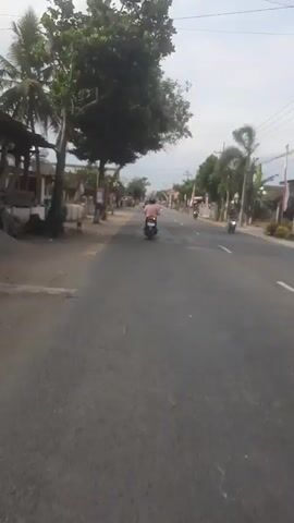 The video shows a paved road with a few motorcycles and people riding them. The road is lined with trees and buildings on both sides. The sky is cloudy. The video appears to be taken from a moving vehicle.