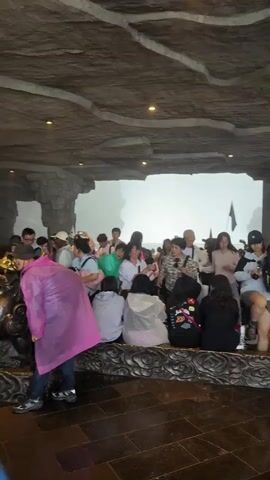 The video shows a group of people gathered in an indoor space, seemingly a covered viewing area or shelter, possibly a stone cave or structure. The walls and ceiling are constructed from grey stone material, giving the impression of a natural cave. Several tourists are milling around, some sitting on a low ledge, while others are standing and walking. Many of the people are wearing rain gear, such as colorful ponchos, suggesting the weather outside may be wet or rainy. Outside the sheltered area, a misty or foggy landscape can be seen through a large opening. There are a few structures or monuments visible further inside the space. The atmosphere appears to be that of a tourist attraction with people exploring and seeking shelter from the elements.