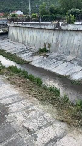 A concrete channelized river flows through a town. The channel has high concrete walls lined with some vegetation. The river appears shallow with a moderate current. The banks of the river are overgrown with grass and some debris. There is a pedestrian walkway with a railing and lampposts along the top of the channel. In the background, there are buildings and trees on a hillside. The sky is overcast.