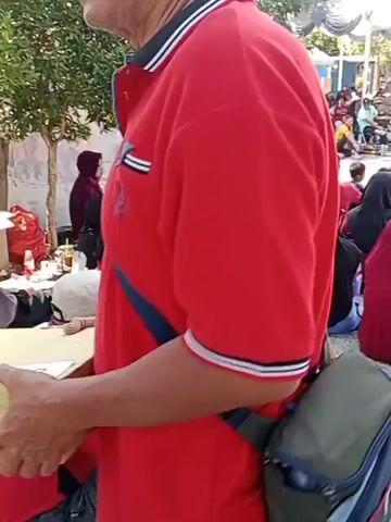 The video shows a person from behind, wearing a red polo shirt with striped cuffs and a blue strap across their chest. They are standing near a table with some papers on it. In the background, there are other people, some sitting and some standing, and a stroller. The environment appears to be outdoors, possibly a market or event, with trees and a tent visible. The person in the foreground is not facing the camera, and their face is not visible. The video is short and provides a brief glimpse of the scene.