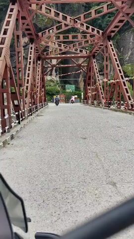 The video shows a point-of-view shot from a vehicle, likely a motorcycle, as it crosses a bridge. The bridge is constructed with red metal trusses and has a paved road surface. On the bridge, two individuals on scooters are traveling in the same direction as the camera. The scooter in front of the camera is ridden by a person wearing dark clothing. The scooter further ahead is ridden by a person wearing pink. The background reveals a steep, rocky mountainside covered in green foliage, suggesting a natural, possibly mountainous, environment. There are flags or banners strung across the bridge and along the sides, adding a festive or decorative element. Signs are visible in the distance. The overall atmosphere appears to be that of travel through a scenic area.