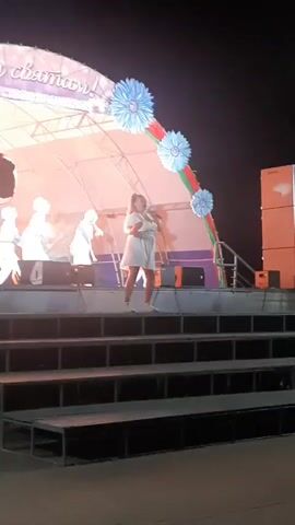 A young woman in a white dress and white sneakers stands on a stage in front of a brightly lit backdrop, holding a microphone and singing. She has blonde hair pulled back in a ponytail. Behind her, on the same stage, are several other performers dressed in white outfits, appearing to be singers or dancers. The stage is decorated with large blue floral accents and red and green ribbons, suggesting a festive or cultural event. The stage is elevated, with a set of dark stairs leading up to it. The lighting is bright, illuminating the performers and the stage area, while the background is dark, indicating it is nighttime. There are speakers visible on the stage.