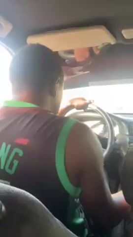 A man is driving a car. He is wearing a black sleeveless shirt with green trim and the word "KING" in green letters on the back. He is wearing an earring in his left ear. He reaches up to adjust the sun visor. The car is moving forward.