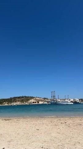 The video shows a sunny beach with a clear blue sky above. The sand is light beige. In the water, several boats are docked, including at least one large white yacht. A landmass in the background features some buildings and tall industrial structures, possibly smokestacks. The water is calm and blue-green. A breakwater of large rocks is visible on the right side of the frame.