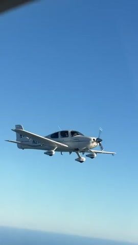 This video shows a small white airplane flying through a clear blue sky. The airplane is in the foreground and is captured in profile, with its wings, propeller, and landing gear clearly visible. There are two people inside the cockpit of the plane. Below the airplane, the ocean is visible, stretching out to the horizon. The camera appears to be filming from another aircraft or from a close proximity to the airplane.