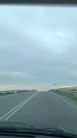 The video shows a point-of-view perspective from inside a vehicle driving on a highway. The road is paved with asphalt and has white lane markings. The sky is overcast with gray clouds. On the right side of the road, there is a guardrail and a yellow warning sign with a black arrow indicating a turn. The road appears to be elevated, possibly crossing over a body of water or a lower land area, as evidenced by the presence of a body of water to the right at the end of the clip. There are no other vehicles or people visible in the frame. The time stamps indicate the video spans about 4 seconds.