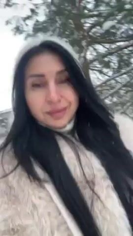 A woman with long black hair and wearing a white fur coat and a white hooded top is walking outside in a snowy environment. The video is shot from a low angle, close to the woman's face. She is looking at the camera, and the background consists of snow-covered trees. The video is slightly shaky, suggesting it might be hand-held.