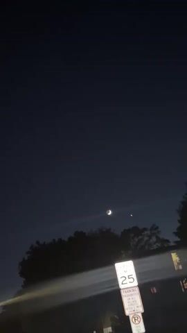 The video is a vertical, nighttime shot of a dark blue sky with a crescent moon and a bright star visible. In the foreground, a speed limit sign reads "25" and below it a parking sign that is partially obscured by motion blur. Trees and the roof of a building are visible at the bottom of the frame. The camera appears to be moving.