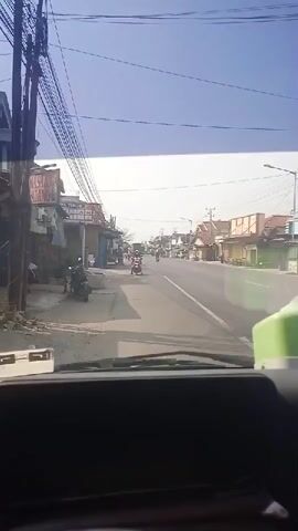 A POV shot from inside a vehicle shows a two-lane road with traffic. On the left side of the road, there are several buildings with storefronts, a few parked motorcycles, and some overhead power lines. On the right side of the road, there are more buildings and storefronts, and some trees. Several motorcycles are riding on the road, some in the foreground and some in the distance. The sky is blue with some wispy clouds.