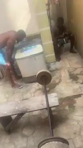 The video shows an outdoor workout area. One man, shirtless and wearing blue shorts and flip-flops, is standing and looking at a barbell. The barbell is lying on a wooden bench and has stone weights attached to each end.  Another shirtless man is sitting in the background using a phone. The area is paved with patterned concrete tiles. A washing machine is visible in the background. Some laundry is hanging in the distance.