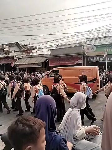 A group of schoolchildren in uniform, appearing to be in a parade or procession, walk down a street. They are wearing light brown uniforms with darker pants or skirts, and many have backpacks. Behind the children, an orange van is visible. The street is lined with buildings and shops, and there are many spectators on the sidewalks. Several motorcycles and scooters are parked or being ridden in the background. The sky is overcast. Some of the spectators are wearing hijabs.