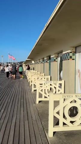 A vertical video shot on a sunny day shows a wooden boardwalk next to a row of beach cabanas. The cabanas have ornate white decorative fences in front of them, each with a name carved into the top railing. The names visible are JAMES, ARLENE DAHL, JEAN, and JEAN MARTIN. A group of people walk along the boardwalk away from the camera, and a couple is seen walking towards the camera. American flags are flying on poles in the background. The beach is visible to the left of the boardwalk, with the ocean and some buildings in the distance.