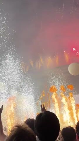 This is a vertical, low-angle video of a live performance, likely a concert. The foreground shows the silhouettes of a crowd with their hands raised. In the midground, on a stage, a golden, life-sized statue of a person stands on a large sphere. Behind the statue, there are pyrotechnics, including sparks and flames erupting from the stage. Laser lights, predominantly red and blue, crisscross the scene from the top of the frame downwards. A large, dark, circular object, possibly a balloon, is visible to the right of the statue. The overall atmosphere is energetic and celebratory, with a focus on dramatic visual effects.