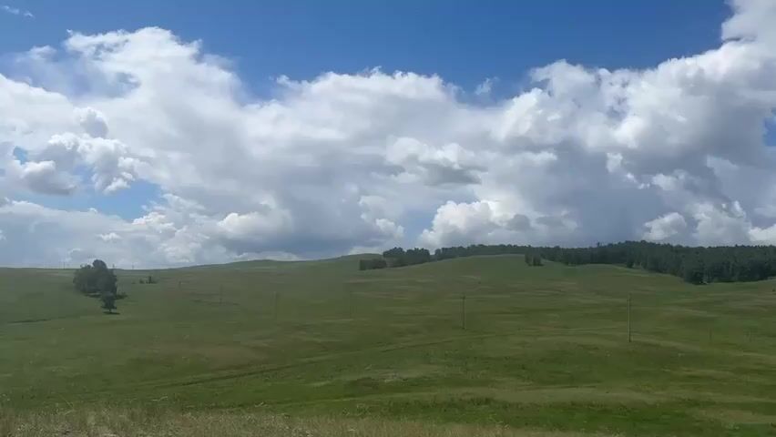 The video shows a panoramic view of a vast, green hilly landscape under a partially cloudy sky. In the foreground, tall grass and a few rocks are visible. As the camera pans to the right, a dirt road winds through the fields, leading to a small village. The village consists of several houses with colorful roofs, scattered across the green expanse. Beyond the village, more rolling hills covered in trees are visible, creating a sense of depth and natural beauty. There are no people or animals visible in the video. The overall impression is one of a peaceful rural setting.