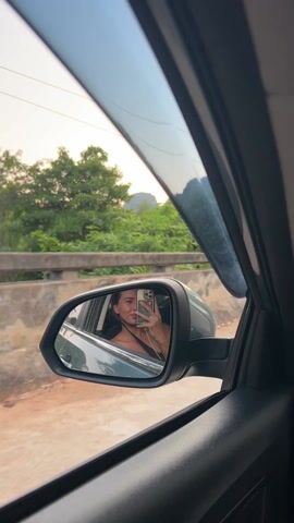 A person sitting in the driver's seat of a car is reflected in the car's side mirror. The person is holding up a cell phone and appears to be taking a selfie. The car is driving on a paved road. Outside the car's windows, there is green vegetation, trees, and a large hill or mountain. There is a speed limit sign visible on the side of the road. The sky is light, and it appears to be early morning or late afternoon.