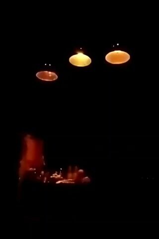 The video is taken in a dimly lit space. Three lights hang from the ceiling, each with a shade that directs the light downwards. Below the lights, in the lower portion of the frame, there is a table or counter displaying various items. A structure, possibly a cabinet or display case, stands to the left of the table. The poor lighting and limited framing make it difficult to discern specific details of the items on the table and the surroundings.