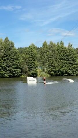 A person is wakeboarding on a lake. They are wearing a helmet and a life vest. There are two ramps in the water. The person is approaching one of the ramps. There are trees in the background. The sky is blue with some clouds.