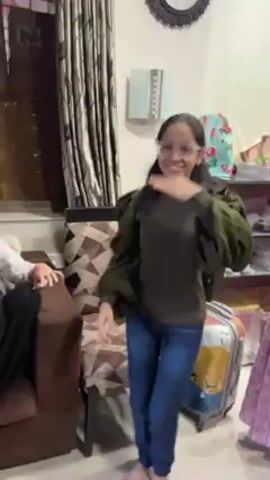 A young girl with glasses and long dark hair dances in front of a brown couch and a suitcase. She wears an olive green top with puffy sleeves and dark blue jeans. She starts by waving her hands, then claps them, then moves them back and forth in a horizontal motion. Behind her, a woman in a pink patterned dress stands to the right. To the left, a person is sitting on the couch. The room appears to be a living area with patterned curtains and a decorative clock on the wall. Music plays in the background.