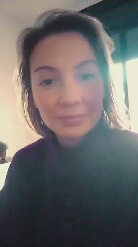 A woman is facing the camera, indoors. She has fair skin, brown hair that is slightly disheveled, and brown eyes. She is wearing a dark purple or black turtleneck sweater. There is a bright window in the background, causing some glare on the camera. A plant is partially visible on a surface in the background. The woman is looking directly at the camera with a slight, ambiguous smile. The video is a close-up shot, focusing on her face.