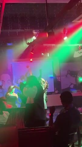 The video shows a lively scene inside a nightclub, with people dancing on stage under colorful, flashing lights. There’s a DJ playing music at the back of the stage. The dancers are dressed casually and moving enthusiastically to the music. A large mural is visible on the wall behind them. The lighting cycles through various colors, creating a vibrant atmosphere. The perspective is from the audience, with heads visible in the foreground.
